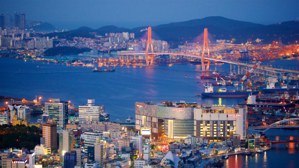 Foto tirada da cidade de Busan, um rio e vários prédios nas margens do rio e uma ponte extravagante que conecta as duas porções de terra