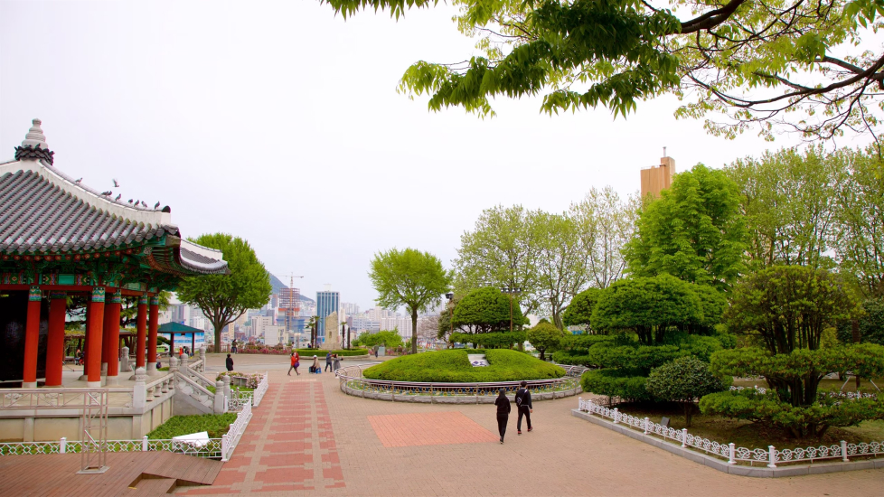 Foto tirada do Parque Yongdusan, mostrando uma construção parecida com um templo e uma grande área para caminhadas, com pequenas áreas de vegetação (grama, arbustos e árvores pequenas), com pessoas algumas poucas indo e vindo.
